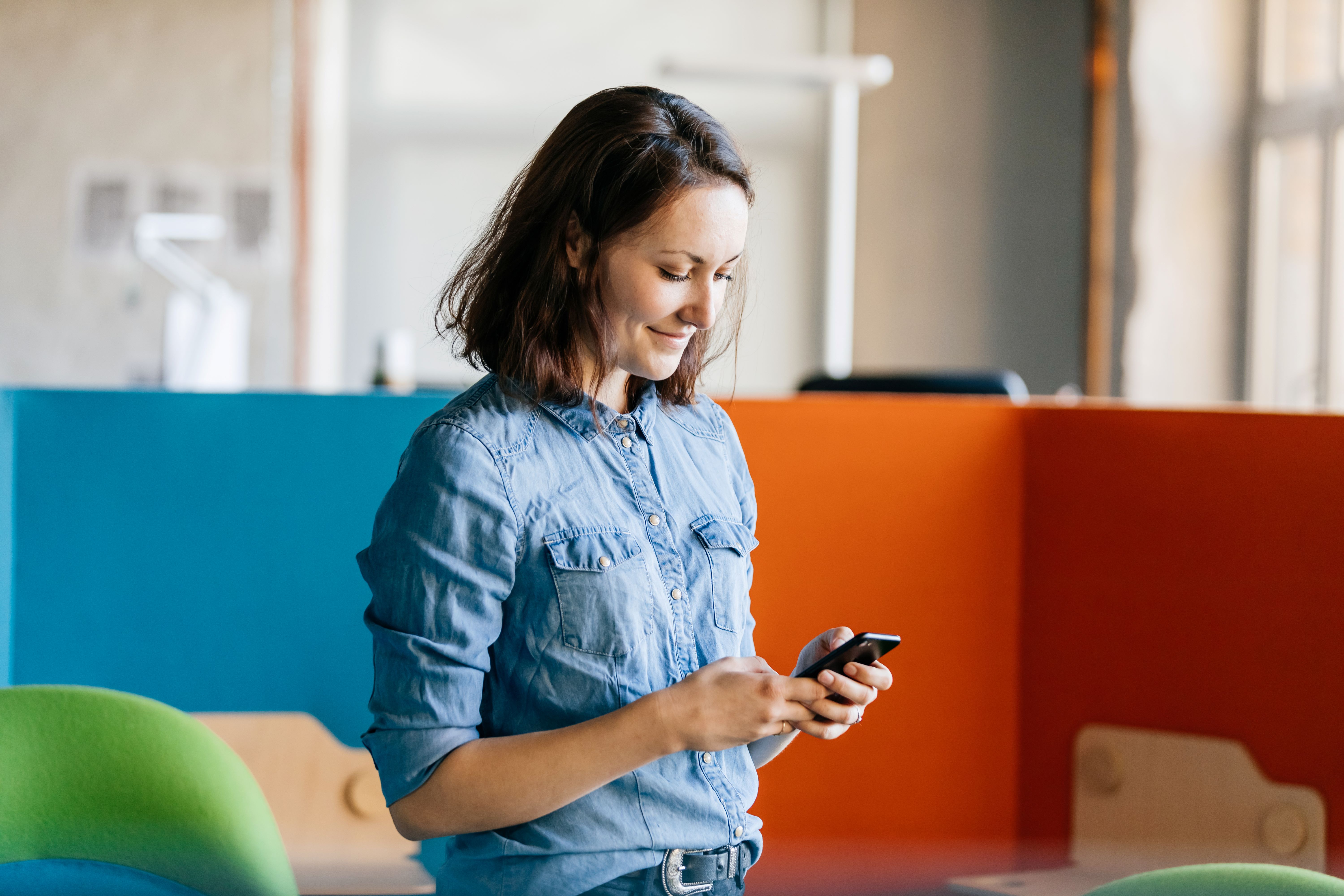 Eine Frau in einem Jeanshemd steht in einem modernen Büro, lächelt und schaut auf ihr Smartphone. Im Hintergrund sind bunte Trennwände und ein Schreibtisch zu sehen.